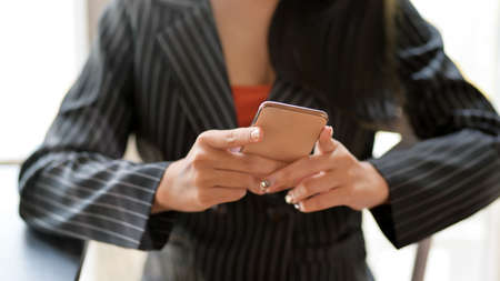 Close up view of female freelancer using smartphone while sitting next to window in dark modern...