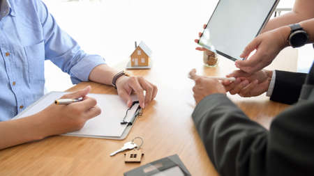 Side view of real estate agent presenting to customer with blank screen tablet, house model and calculator on wooden deskの写真素材
