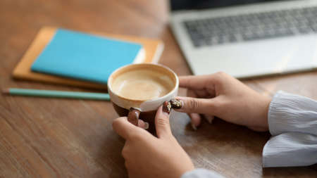 Close up view of a woman holding coffee cup in comfortable workplace with laptop and stationeryの写真素材
