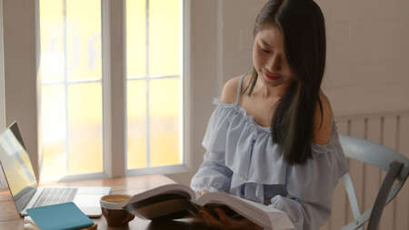 Cropped shot of a girl reading book while siting next to window in comfortable room with laptop and stationeryの写真素材