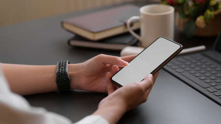 Close up view of a female using blank screen smartphone on black desk with notebooks and coffee cup...
