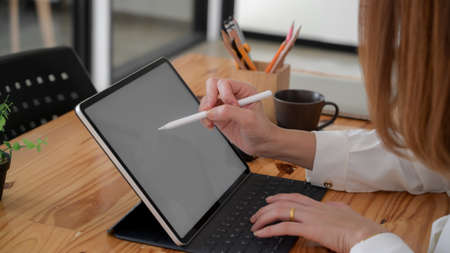 Side view of female freelancer drawing on blank screen tablet with stylus and other office supplies...