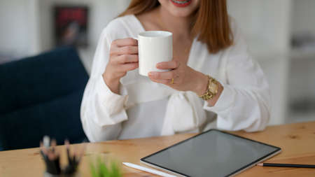 Close up view of female freelancer holding a coffee cup while working with digital tablet in simple co working spaceの写真素材