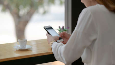 Cropped shot of a woman texting on  smartphone while sitting next to window in simple coffee shop