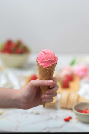 Cropped shot of a girl holding strawberry ice-cream cone with blurred strawberry and topping on...