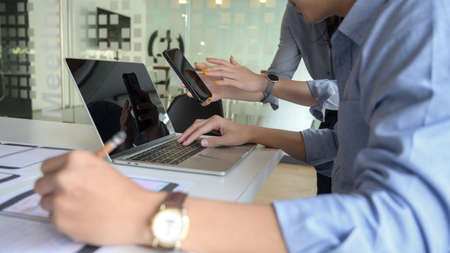 Side view of young male UI developer working on laptop on white desk while consulting with co worker in simple office roomの写真素材