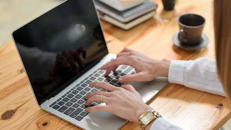Cropped shot of a woman typing on laptop on wooden desk with coffee cup and office supplies