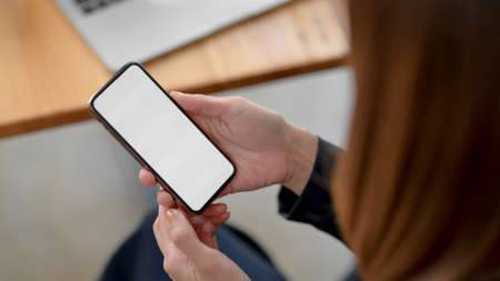 Cropped shot of businesswoman using blank screen smartphone with laptop on wooden desk while sitting...