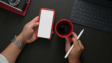Overhead shot of male photographer holding mock up smartphone while working in stylish workplace with stylus, tablet and camera on black desk   の写真素材