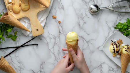 Overhead shot of a girl holding vanilla ice-cream cone on marble desk with topping tray and...