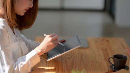 Side view of female freelancer working on digital tablet with stylus and coffee cup on wooden table...
