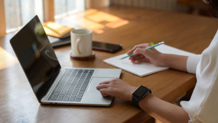 Cropped shot of young college girl working on laptop while writing on notebook in simple workspace 