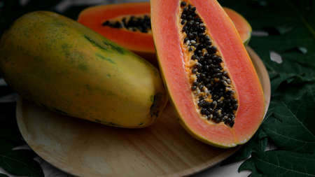 Cropped shot of whole and halved sweet papaya on wooden plate and papaya leaves on marble table 