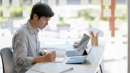 Side  view of young male entrepreneurs focusing on his work while using laptop, tablet and other...