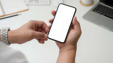 Cropped shot of businessman holding blank screen smartphone on white desk with laptop and office...