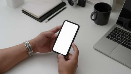 Cropped shot of male holding blank screen smartphone on white desk with laptop and office supplies in minimal workspaceの写真素材