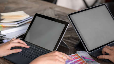 Close up view of two young designers consulting for choosing colour on colour swatch with mock-up tablets on wooden deskの写真素材