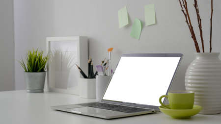 Cropped shot of stylish workplace with blank screen laptop, stationery, green cup and decorations on...