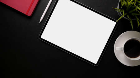 Top view of stylish workspace with blank screen tablet, office supplies and coffee cup on black...