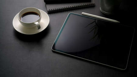 Cropped shot of dark modern workspace with digital tablet, office supplies and coffee cup on black...