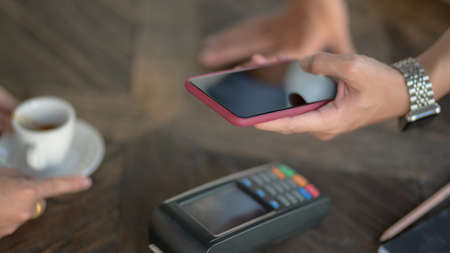 Cropped shot of male customer using smartphone to online payment on wooden desk in coffee shop 