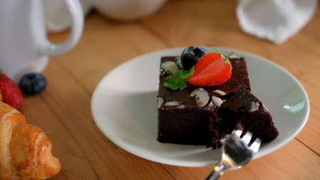Close up view of a girl eating chocolate brownie on white plate with mint leaf, strawberry and...