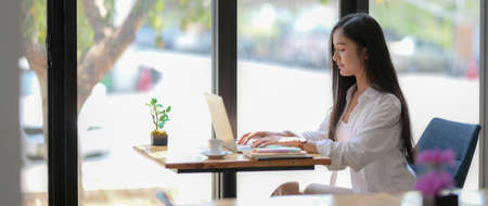 Side view of young female university student typing on laptop on wooden table in coffee shopの写真素材