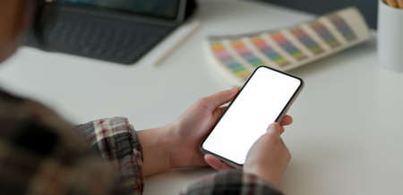 Cropped shot of female designer holding mock-up smartphone while working with tablet and designer supplies on white tableの写真素材