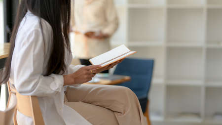 Side view of female university student reading a book while sitting on wooden chair in co-working...