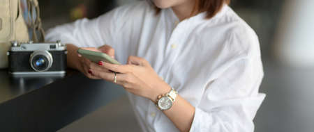 Close up view of young female photographer working with smartphone and camera while sitting at...