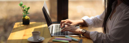 Side view of young college student typing on laptop with stationery and coffee cup on wooden desk in...