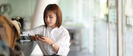 Close up view of young female photographer using smartphone while waiting for customer in glass wall officeの写真素材