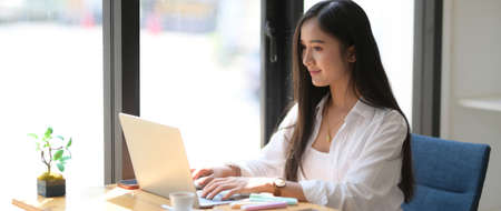 Cropped shot of young college student focusing on her assignment with laptop on wooden desk in...