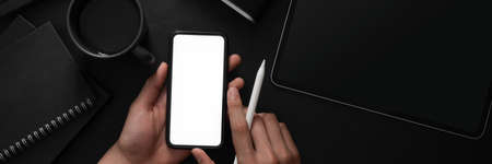 Overhead shot of a man holding blank screen smartphone in dark modern concept workspace with other...