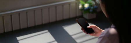 Cropped shot of female freelancer touching on smartphone while sitting at dark modern workspaceの写真素材