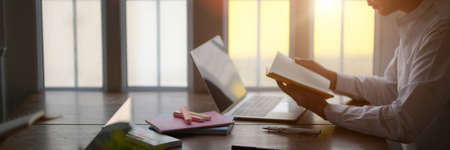 Side view of young male university student preparing for exam with laptop and his notebook on wooden desk in libraryの写真素材