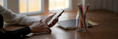 Side view of female freelancer holding blank screen smartphone on wooden table with supplies and...