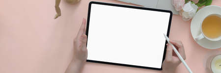 Overhead shot of female university student doing homework with blank screen tablet on pink table...