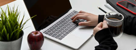 Close-up view of woman typing on laptop computer while drinking a cup of coffee in her workspace