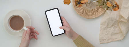 Top view of female freelancer using blank screen smartphone on breakfast table with decorations and drinking morning coffeeの写真素材