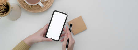 Overhead shot of female freelancer using mock-up smartphone to contact customer on white table with...