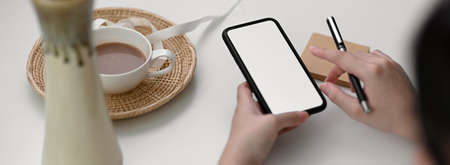 Cropped shot of female freelancer using mock-up smartphone to contact customer on white table with...