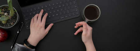 Overhead shot of female entrepreneur working on digital device on black table and drinking hot...