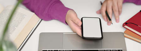 Top view of female college student looking on mock-up smartphone while doing assignment with laptop and books on white tableの写真素材