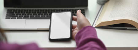 Close up view of college student using smartphone to relax while doing assignment with laptop on white tableの写真素材