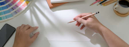 Top view of male artist drawing on sketch paper on white worktable with smartphone and designer...