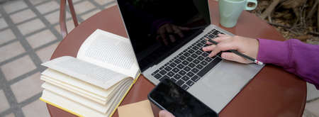 Cropped shot of female university student doing her assignment with laptop, smartphone and book on...