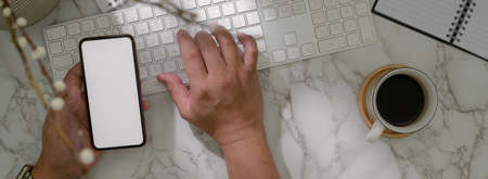 Overhead shot of male entrepreneur working with computer keyboard, mock-up smartphone on marble...