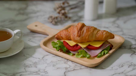 Close up view of breakfast meal with croissant sandwich ham, cheese on wooden plate and tea cup on marble desk の写真素材