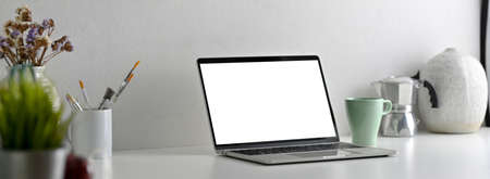 Side view of modern office desk with blank screen laptop, painting tools and decorations on white...